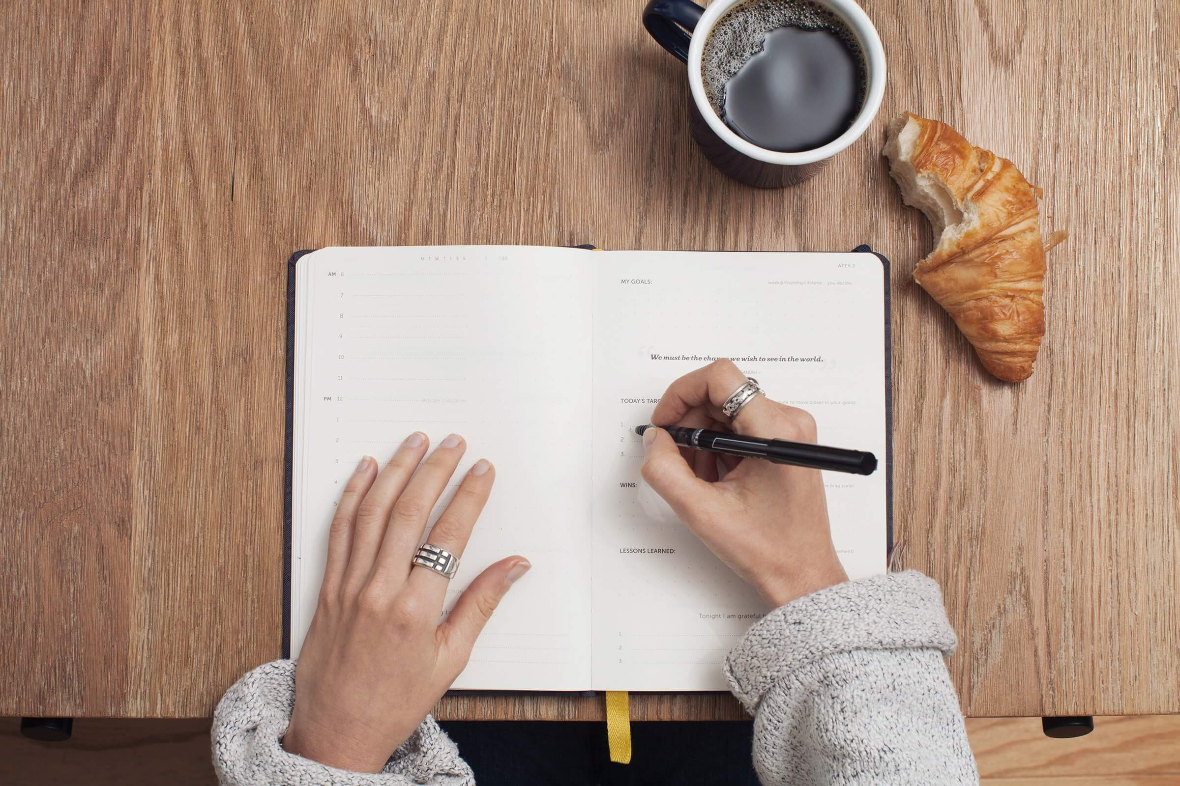 Overhead photo of woman writing in notepad with croissant and cup of coffee beside the notepad